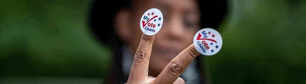 Photo of person holding up two fingers with My Vote Counts stickers on fingertips.
