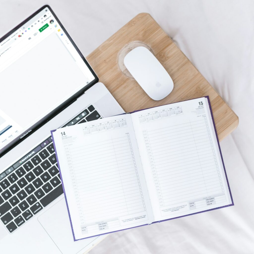 Laptop and mouse on a wooden desk, with an open notebook on top of the keyboard