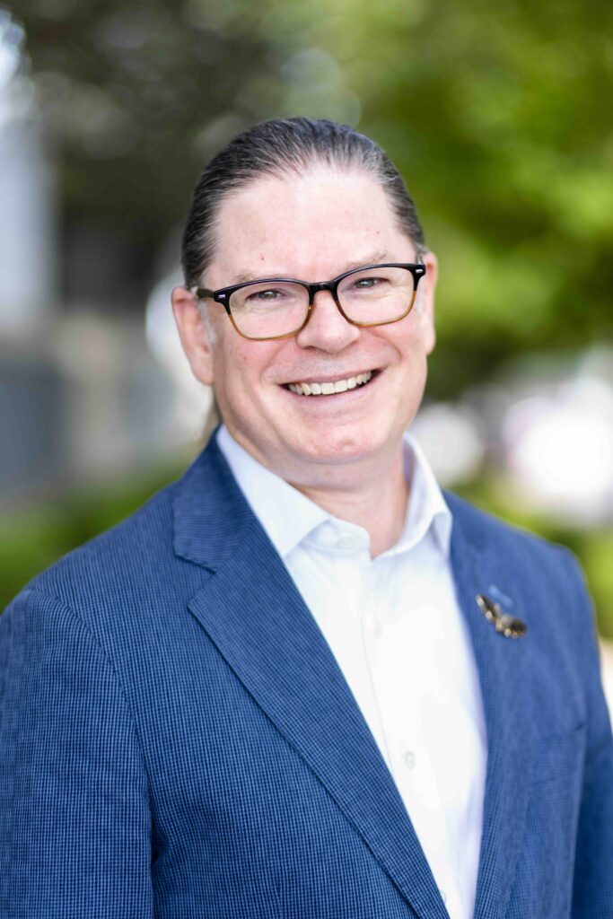 Man smiling, wearing brown glasses, a shirt, and a blue blazer, outdoors with a green background.