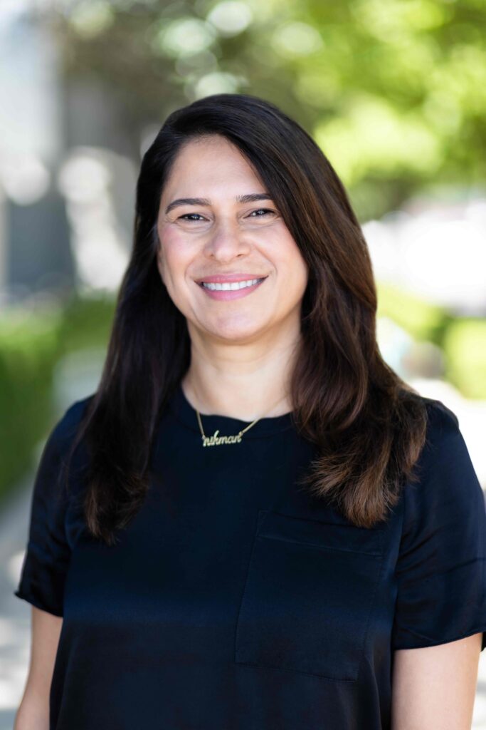 Woman smiling, with long brown hair, wearing a navy blue blouse, outdoors with a green background.