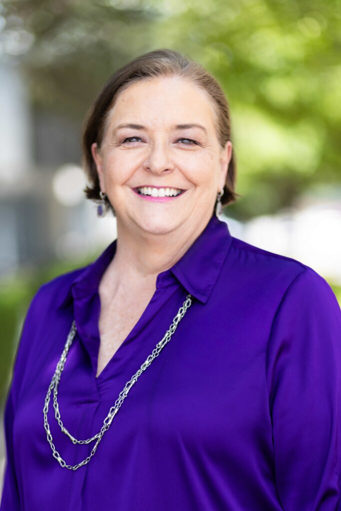 Woman with short hair, smiling, wearing a purple blouse with a long silver necklace, outdoors with a green background